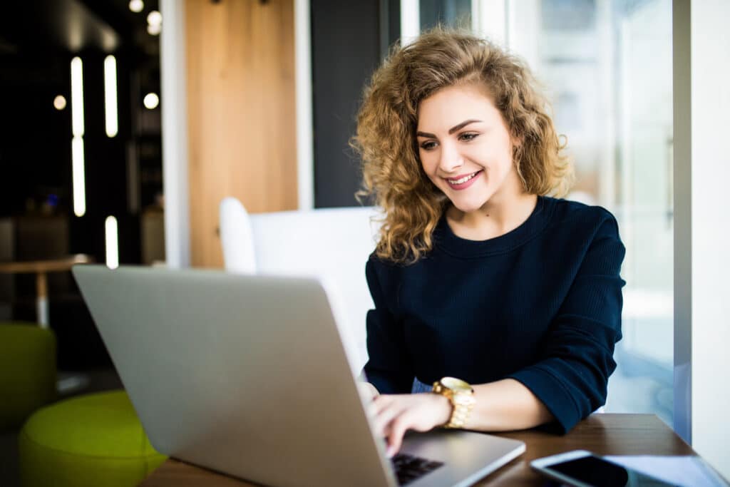 Happy curly young woman use laptop in modern place with bright windows.