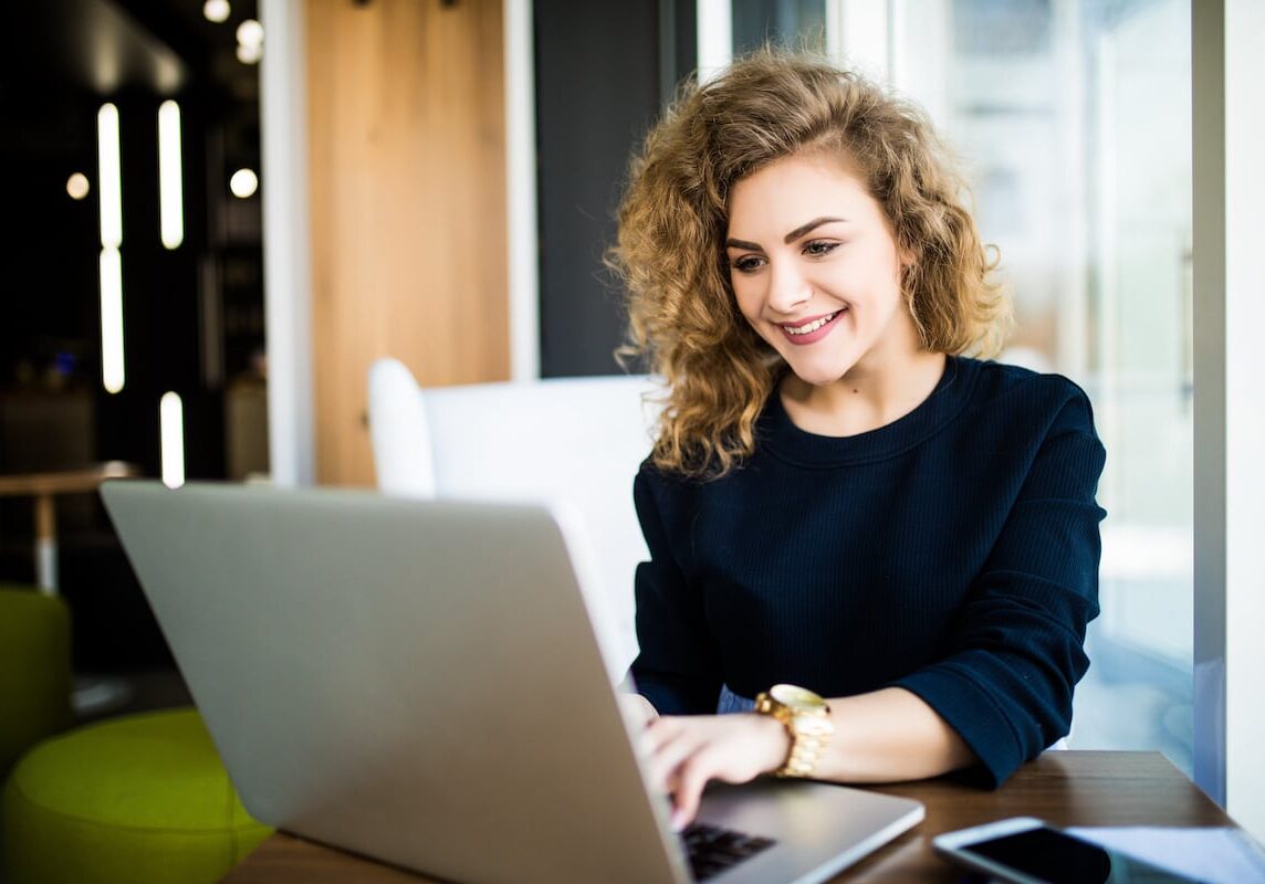 Happy curly young woman use laptop in modern place with bright windows.
