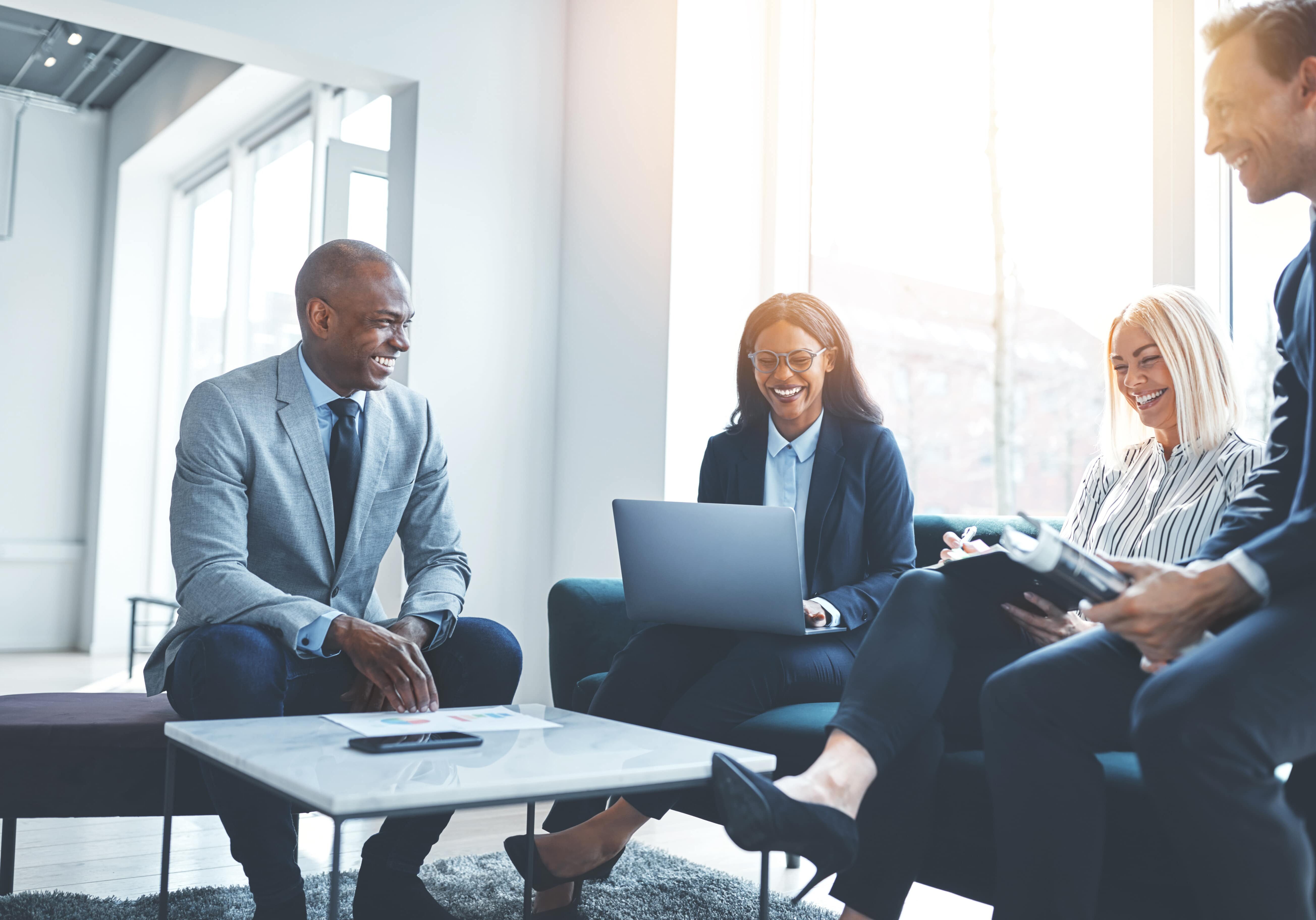 Diverse group of smiling businesspeople talking together while sitting on a sofa in a bright modern office