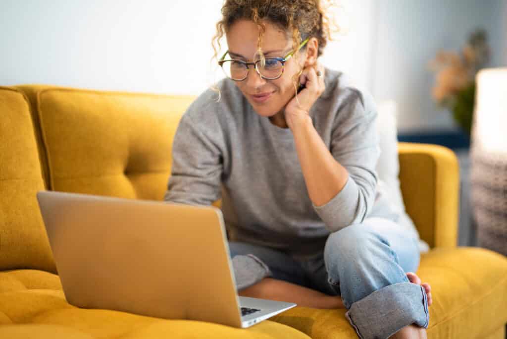 Female multifamily marketer sitting on a yellow couch using laptop and internet connection and smile.